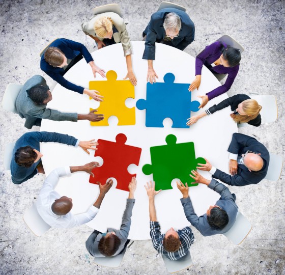 Overhead view of people sitting at a round table, all reaching out to touch large puzzle pieces